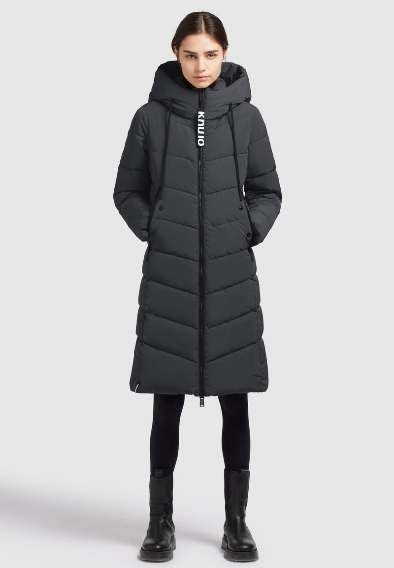 Mustal puffer-jakk pikendatud pikkus, kapuutsiga, ševronmuster, esi-zipper sulgemine, nööriga, ja nähtav logo detail zipperi peal.