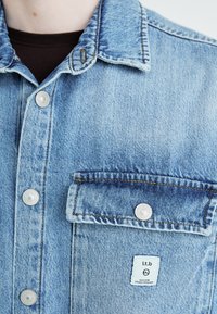 Giacca di jeans blu chiaro con colletto a punta, bottoni a pressione argentati, una tasca sul petto con etichetta ricamata e cuciture gialle a contrasto.