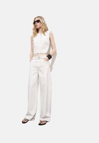 Top senza maniche bianco corto abbinato a pantaloni bianchi a gamba larga. Gli accessori includono una cintura con borchie e diversi bracciali neri. Occhiali da sole scuri.