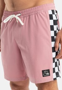 Los pantalones de baño rosas cuentan con un panel lateral a cuadros en blanco y negro, una cinturilla elástica con un cordón blanco y un parche con el logo en el dobladillo.