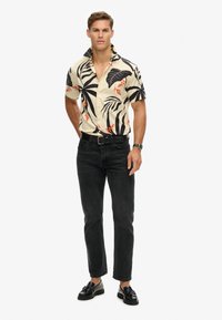 Camisa de manga corta con botones que presenta una base crema con patrones de hojas tropicales en negro y naranja. Combinada con jeans negros y zapatos.