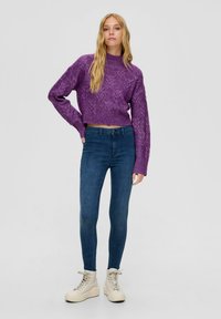 Zkrácený fialový svetr s pleteným vzorem a vysokým límcem, spárovaný s tmavě modrými úzkými džíny a béžovými vysokými teniskami.