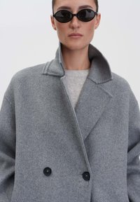 Vrouw met zwarte ovale zonnebril en een grijze wollen jas met een brede kraag en twee zwarte knopen boven een licht truitje.