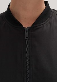 Czarna kurtka bomberka z zamkiem błyskawicznym i ściągaczem przy kołnierzu. Materiał wydaje się gładki; wzór obejmuje minimalną liczbę szwów dla smukłego wyglądu.