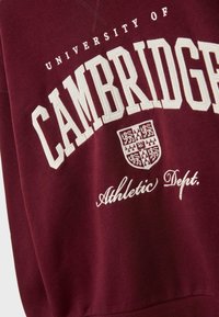 Bordeaux stof med hvid broderet tekst "University of Cambridge Athletic Dept." og et detaljeret broderet våbenskjold i midten.