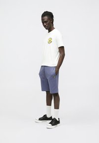 Valkoinen puuvillainen t-paita, jossa keltainen kuvio; violetit shortsit, joissa nyöritys; mustat lenkkarit ja valkoiset sukat. Yksinkertainen muotoilu ja rento istuvuus.