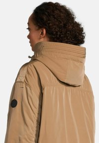 Beige jas met capuchon, voorzien van een geborduurde kraag en schouderaccenten, zachte textuur en een ronde logo patch op de mouw.