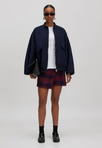 Marineblaue Bomberjacke mit Reißverschluss, kombiniert mit einem rot-navy karierten Minirock. Schwarze Sonnenbrille und eine Lederhandtasche vervollständigen den Look.