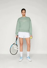 Minta zelena majica s bijelim tekstom, bijele kratke hlače, bijele anklet čarape, plave i bijele teniske cipele, drži tenisku reket i lopticu.