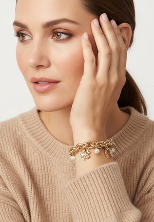 Gouden kettingarmband met parelaccenten en bedeldetails, gedragen aan een hand die tegen de wang rust. Zachte beige trui textuur op de achtergrond.