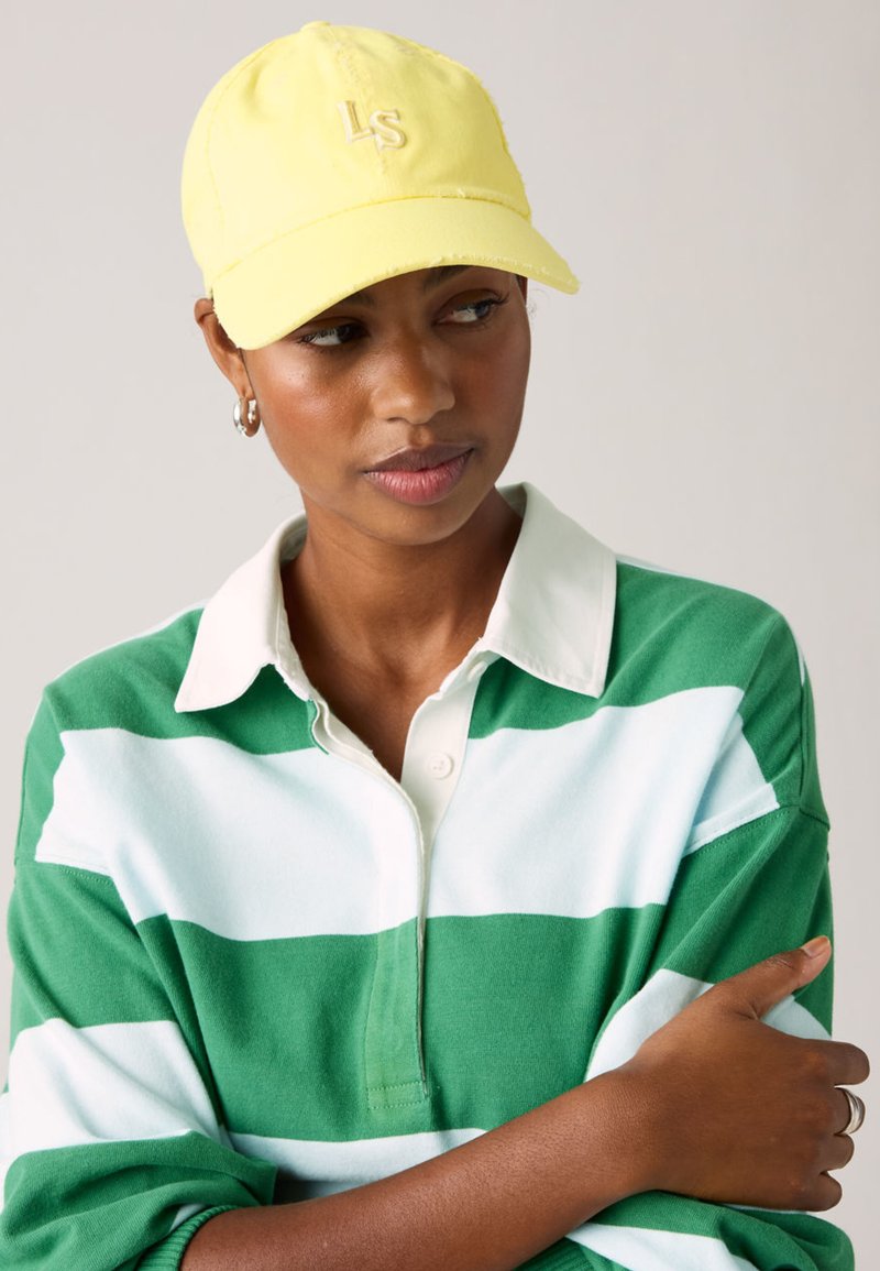 Γυναίκα που φορά κίτρινο καπέλο με το λογότυπο "LS", πράσινο και λευκό ριγέ πουκάμισο με γιακά, κοιτάζει προς τα πλάγι α με σταυρωμένα τα χέρια.