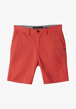 Garçon portant un polo noir, un short corail, des chaussettes blanches à hauteur de la cheville et des baskets Converse noires, debout devant un fond blanc.