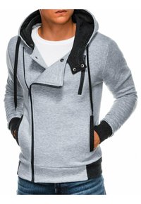 Sweat à capuche zippé gris avec une capuche noire contrastante, des poches latérales et un design unique du col. Confectionné dans un tissu doux avec des poignets côtelés.