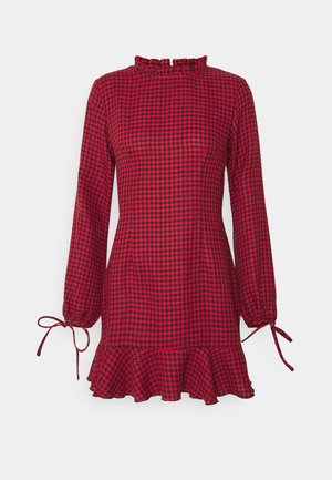 Robe à manches longues à motif pied-de-poule rouge et noire avec un ourlet à volants, un col montant et des poignets noués sur fond blanc.
