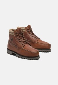 Bottes en cuir marron avec un col rembourré, œillets en métal et une semelle en caoutchouc robuste. Présente une couture décorative et un logo sur le côté.