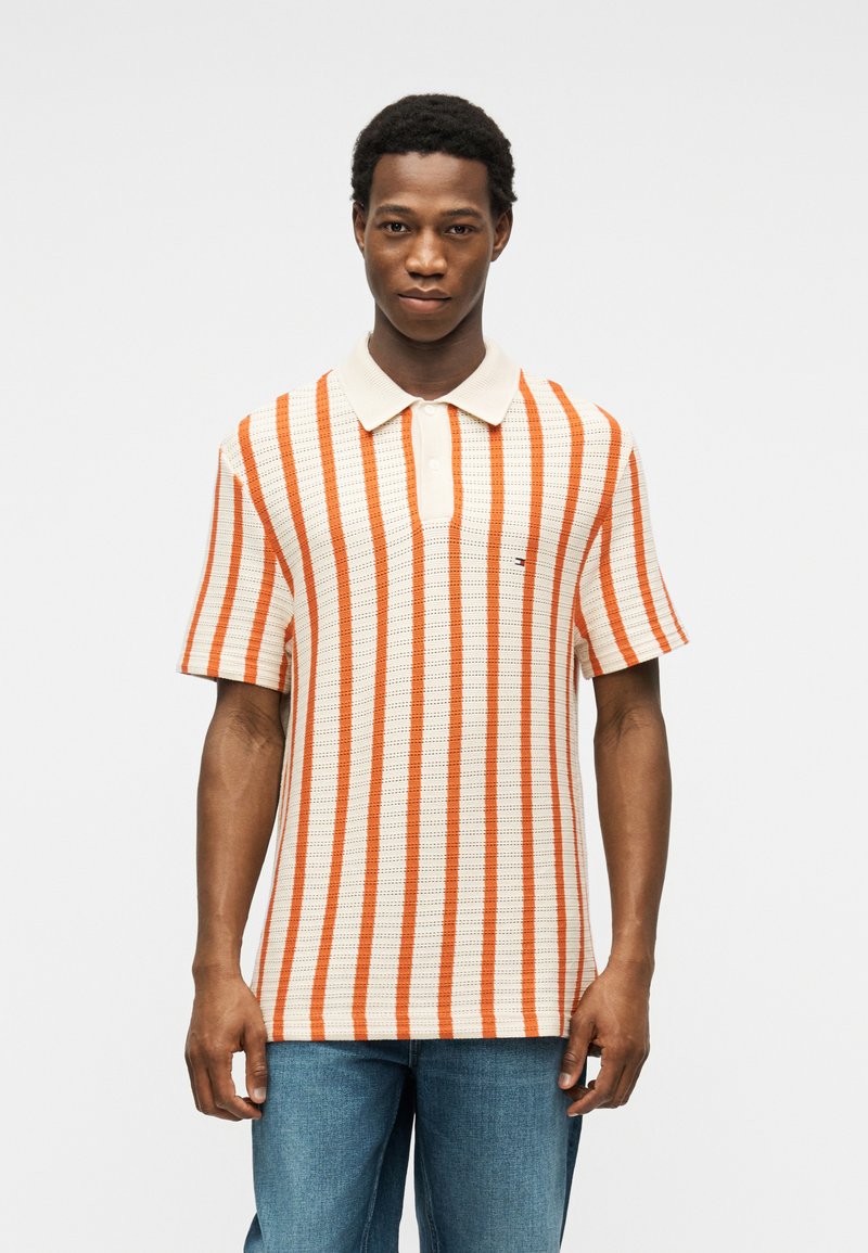Homme portant un polo beige à manches courtes avec des rayures verticales orange et un jean bleu, debout devant un fond blanc uni.