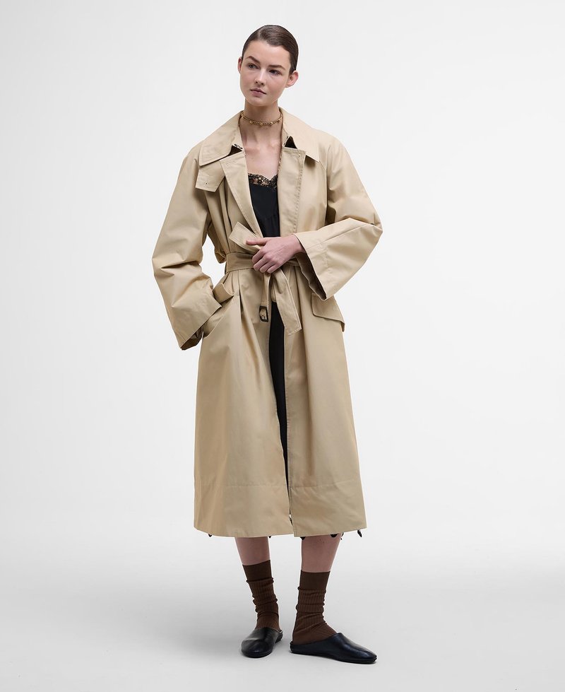 Beige, wasserdichter Trenchcoat mit großem Kragen, weiten Ärmeln und einem Bindegürtel. Das Model trägt ein schwarzes Kleid und braune strukturierte Socken.