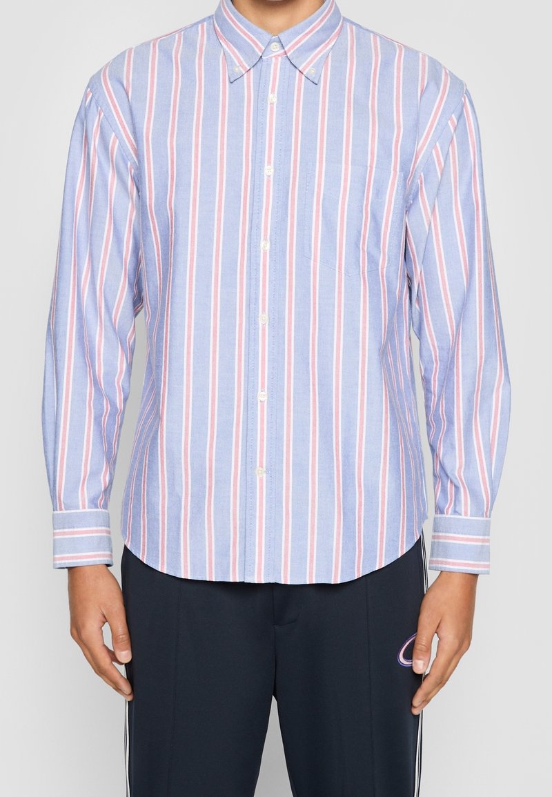 Homme portant une chemise bleu clair boutonnée avec des rayures verticales roses et blanches, associée à un pantalon foncé avec un petit logo.