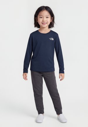 Bambino piccolo in piedi che sorride, indossa una maglia a maniche lunghe blu navy con il logo The North Face, pantaloni grigio scuro e scarpe bianche slip-on.