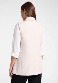 Beige ärmelloser Blazer mit taillierter Passform, glatter Textur und spitzem Kragen, kombiniert mit einem weißen Hemd und hochgerollten Ärmeln.