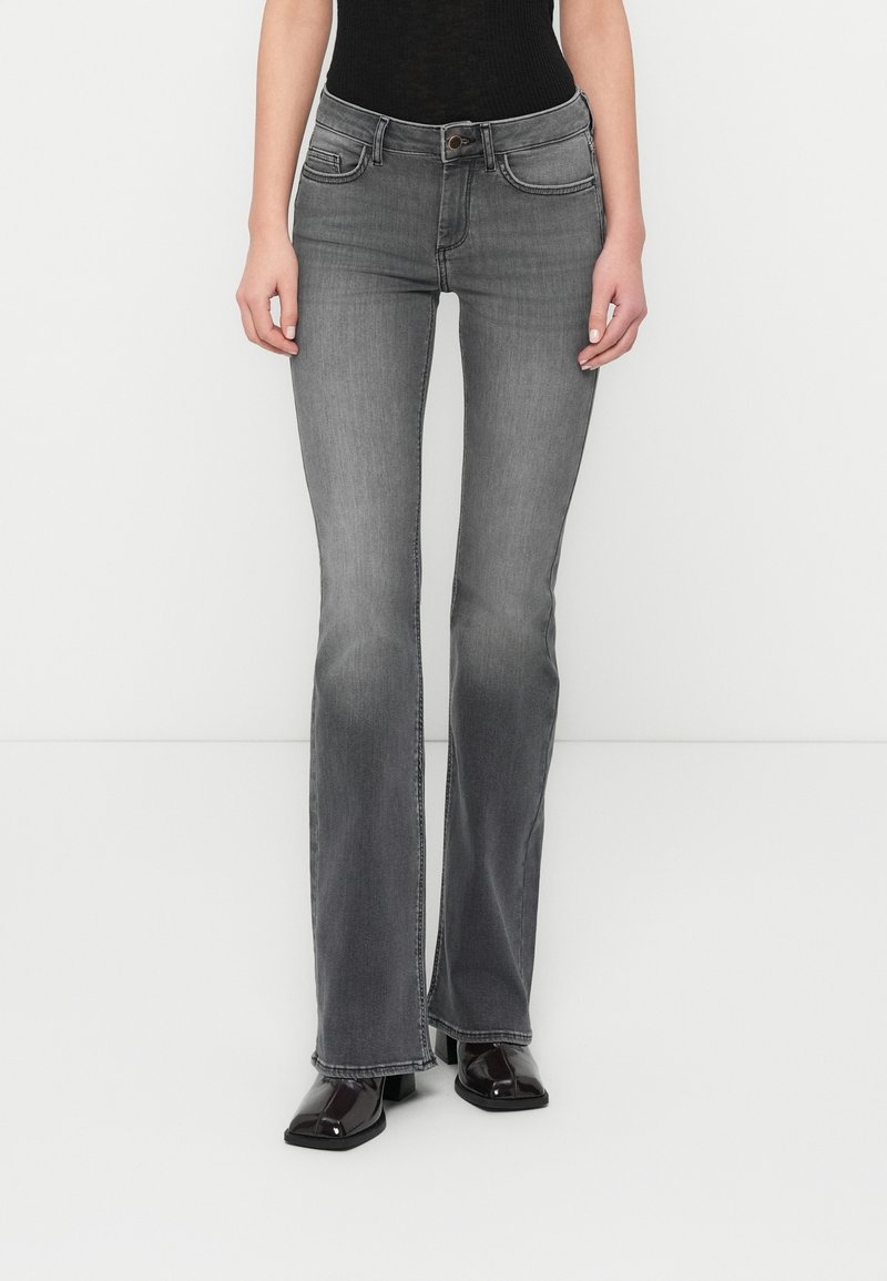 Femme portant un jean gris délavé coupe bootcut et des chaussures à talons noires brillantes à bout carré, debout sur un fond blanc.