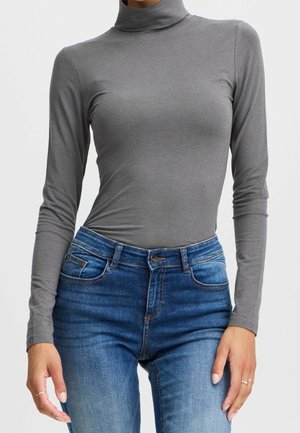 Vrouw die een nauwsluitend grijs coltrui-shirt met lange mouwen en een blauwe spijkerbroek draagt, staat met haar handen ontspannen langs haar zij.