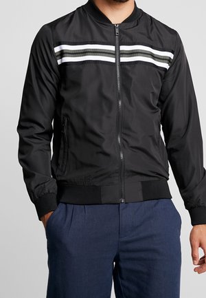 Homme portant une veste noire à fermeture éclair avec des rayures horizontales blanches et grises sur la poitrine, associée à un pantalon bleu foncé.