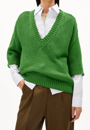 Green knit sweater with a V-neck, loose fit, short sleeves, and ribbed hem, layered over a white shirt and brown trousers.