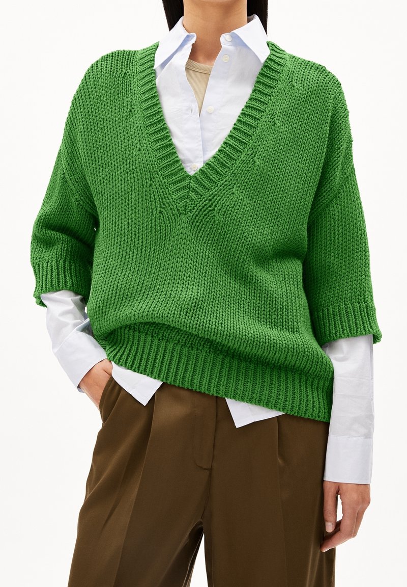 Pull en tricot vert avec un col en V, coupe ample, manches courtes et ourlet côtelé, superposé sur une chemise blanche et un pantalon marron.