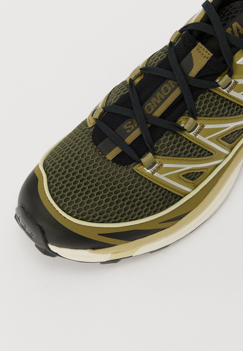 Grüner und schwarzer Sportschuh mit einem Obermaterial aus Mesh, verstärkter Zehenkappe, gepolstertem Kragen und reflektierenden Details auf einer geschwungenen Sohle.