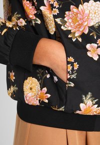 Kurtka bomber z kwiatowym wzorem na czarnym tle, ozdobiona różowymi i złotymi kwiatami, z prążkowanymi mankietami i bocznymi kieszeniami. Miękka tkanina o gładkim wykończeniu.
