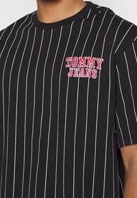 Black short-sleeve t-shirt with white vertical stripes and a red embroidered "TOMMY JEANS" logo on the left chest. Cotton material.