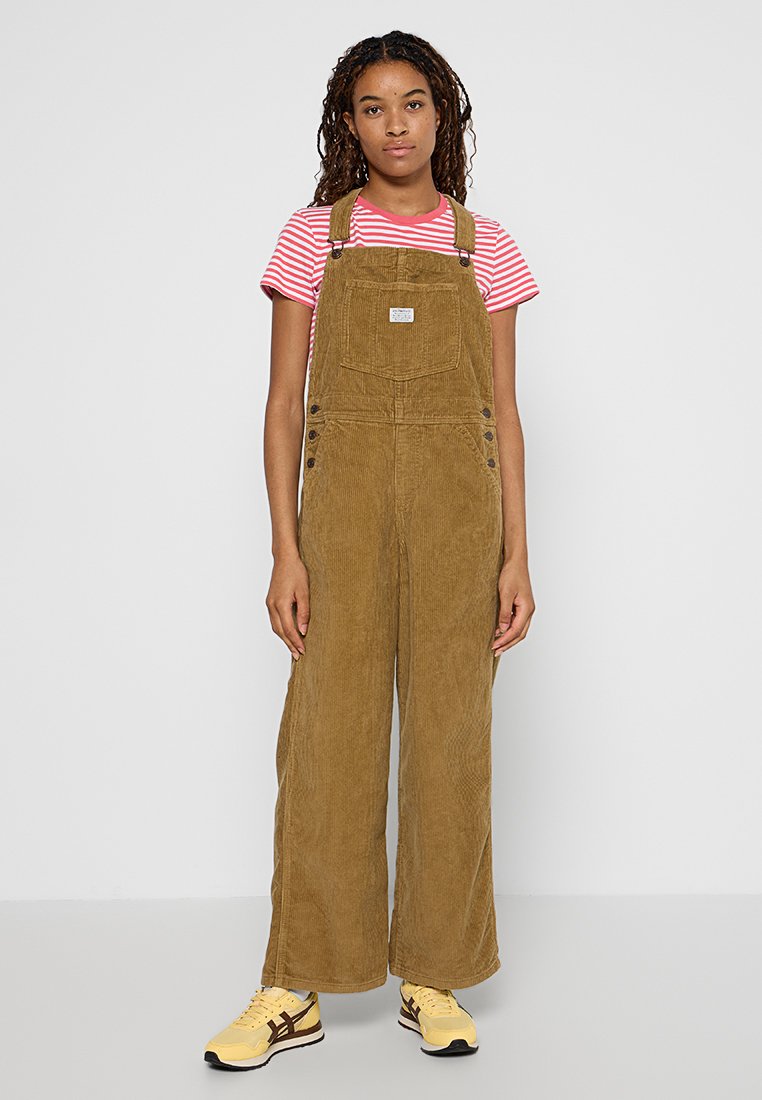 Salopette en velours côtelé marron avec des jambes larges, bretelles réglables et boutons-pression. T-shirt rayé rouge et blanc en dessous. Baskets jaunes.
