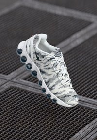 Zapatilla deportiva blanca con un patrón de camuflaje gris, suela redondeada con acentos en azul verdoso, parte delantera con cordones y un cuello acolchado para mayor soporte.