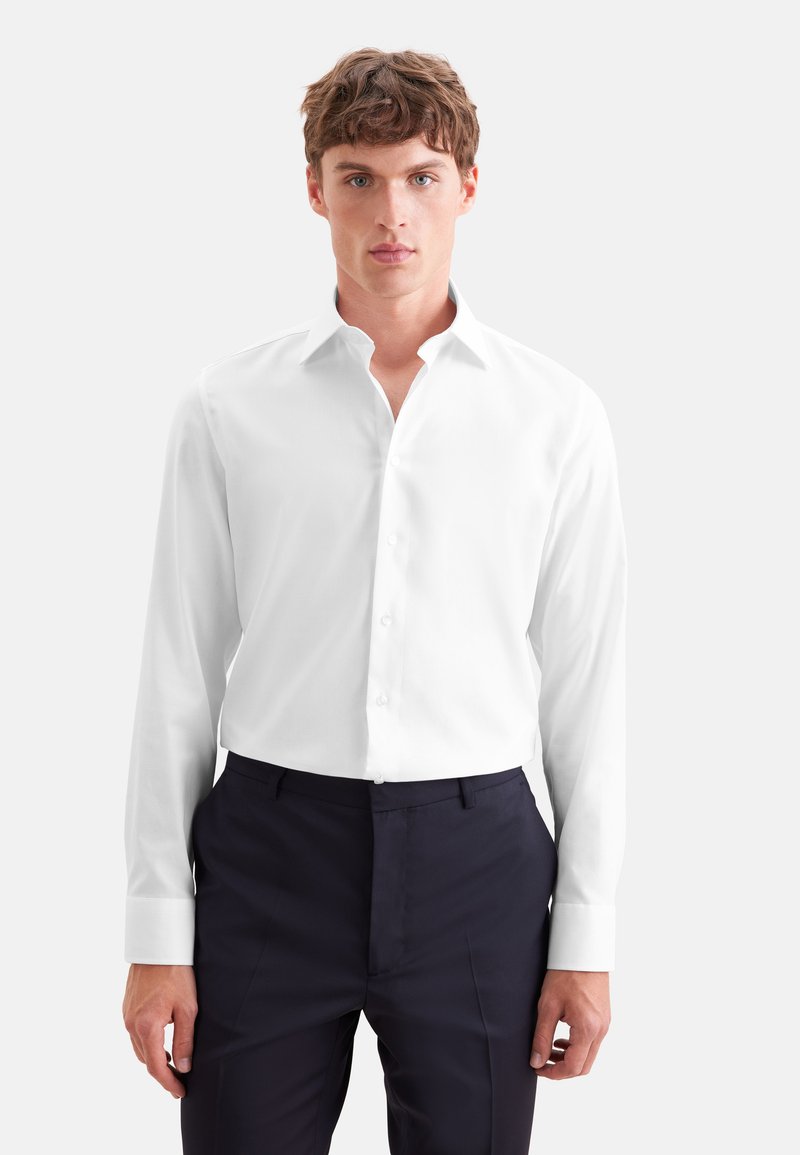 Jeune homme aux cheveux bruns courts portant une chemise blanche à manches longues boutonnée et un pantalon bleu marine foncé, debout devant un fond clair uni.