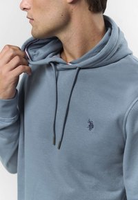 Svetlo modra jopa s kapuco iz bombažne mešanice, z žepom kangaroo spredaj, kapuco z vrvico in subtilnim logotipom, izvezenim na prsih.