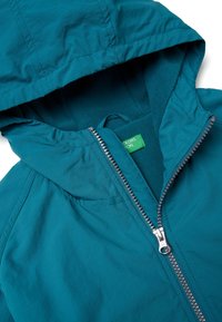 Tyrkysová bunda s kapucí a fleecovým zateplením, která má stříbrný zip a štítek. Tkanina je hladká a matná.