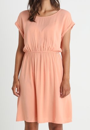 Kobieta ubrana w brzoskwiniową sukienkę o długości do kolan, z krótkimi rękawami, z marszczoną elastyczną talią i okrągłym dekoltem.