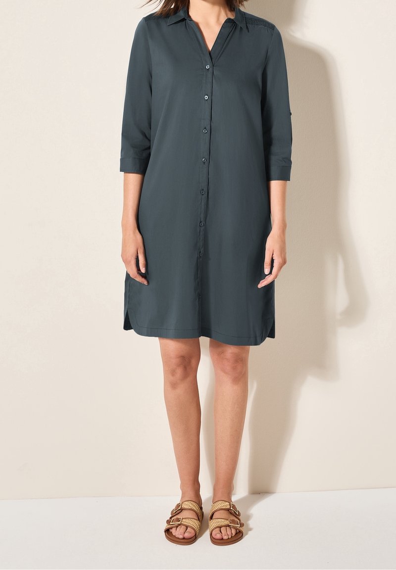 Femme portant une robe-chemise bleu sarcelle foncé à boutons avec des manches trois-quarts et des sandales à double bride beige, debout contre un mur clair.