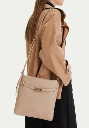 Donna con giacca beige e gonna nera che porta una borsa a tracolla in pelle beige con fibbia dorata e logo LASOCKI.