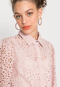 Camisa rosa claro com recortes florais bordados, colarinho, fecho de botão e mangas curtas bufantes. Textura suave com um design delicado.