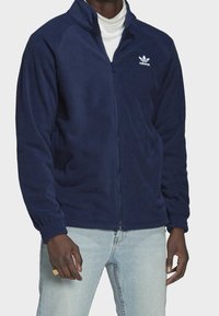 Mörkblå fleecetröja med hög krage, dragkedja fram, elastiska muddar och den vita Adidas-logotypen på vänster bröst.