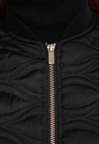 Czarna pikowana kurtka z falistymi wzorami tekstury, złotym suwakiem i wysokim kołnierzem. Materiał jest gładki o miękkim wykończeniu.