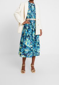 Robe fleurie bleue et jaune avec une taille cintrée, assortie d'un blazer crème. Elle présente une jupe longueur genoux et des talons jaunes à lanières.