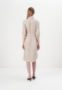 Robe chemise en lin beige avec une ceinture, manches longues retroussées, empiècement au dos et fentes sur les côtés pour faciliter les mouvements.