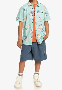 Kurzärmliges mintgrünes Button-Up mit floralen Muster, orange T-Shirt, blaue Shorts und weiße Sneakers mit mehrfarbigen gestreiften Socken.