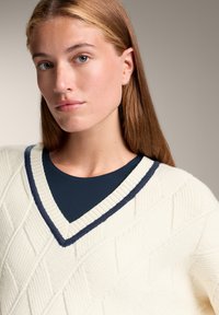 Cremfarbener gestrickter Pullover mit einem Rautenmuster, ausgestattet mit einem marineblauen V-Ausschnitt. Über einem dunkelblauen Oberteil getragen, wodurch Textur und Layering zur Geltung kommen.