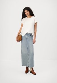 Top bianco a maniche corte, gonna maxi in denim azzurro chiaro, stivali con tacco marroni con dettagli bianchi e una piccola borsa marrone. Tessuto semplice.
