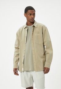 Camisa beige de botones con un bolsillo delantero, tejido ligero y botones plateados, superpuesta sobre una camiseta verde claro y pantalones cortos beige.
