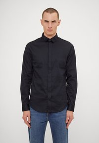 Camisa negra de manga larga con cuello abotonado, hecha de una tela suave. Presenta un logo sutil y puños en contraste, combinada con jeans de color azul claro.